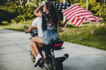 Pareja joven sosteniendo la bandera estadounidense mientras conduce una motocicleta en la carretera rural, Krabi, Tailandia, vista trasera - foto de stock