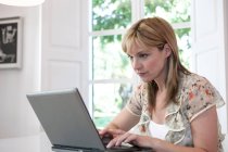 Mulher madura digitando no laptop por janela — Fotografia de Stock
