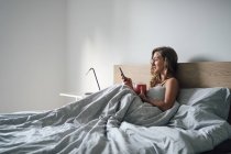 Молода жінка сидить у ліжку зі смартфоном — стокове фото