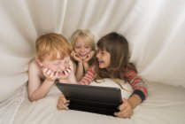 Діти лежать в ліжку з цифровим планшетом і сміються — стокове фото