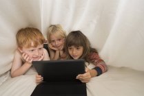 Діти лежать у ліжку і дивляться на цифровий планшет — стокове фото
