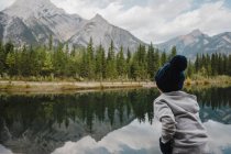 Boy looking at reflection of mountain and trees in lake, Canmore, Canadá, América do Norte — Fotografia de Stock