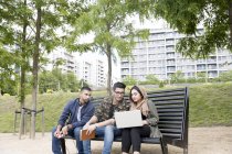 Três amigos sentados no banco no parque e olhando para o laptop — Fotografia de Stock