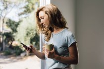 Junge Frau an Terrassentür schaut auf Smartphone — Stockfoto