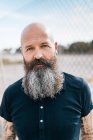 Portrait of mature male hipster with grey beard in front of wire fence — Stock Photo