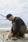 Accovacciato giovane uomo preparare amo da pesca sulla spiaggia — Foto stock