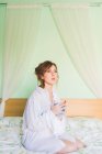 Молода жінка на колінах на ліжку тримає чашку кави і дивиться — стокове фото