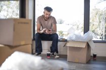 Mann sitzt in unmöblierter Wohnung umgeben von Pappkartons und benutzt Smartphone — Stockfoto