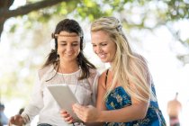 Zwei junge Freundinnen sitzen mit digitalem Tablet unter einem Baum und hören Kopfhörer — Stockfoto