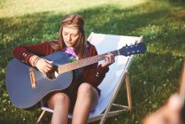 Junge Boho-Frau sitzt auf Liegestuhl und spielt Akustikgitarre beim Festival — Stockfoto