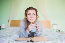 Молода жінка, лежачи на ліжку проведення чашку кави і дивлячись — стокове фото
