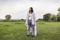 Jeune femme enceinte debout dans le champ, tenant l'estomac, couverture autour des épaules, expression coûteuse — Photo de stock
