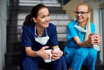 Два хирурга делают перерыв, сидят на ступеньках, держат чашки кофе, улыбаются — стоковое фото