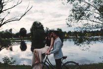 Romántica pareja joven en bicicleta mirándose el uno al otro junto al lago al atardecer - foto de stock