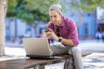 Jovem ao ar livre, sentado no banco, usando laptop, segurando taça de café takeaway — Fotografia de Stock