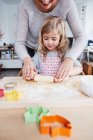 Мати допомагає доньці розгорнути тісто для печива на кухонному столі, середня секція — стокове фото
