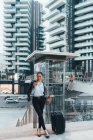 Femme d'affaires avec valise à roulettes utilisant un smartphone à l'extérieur — Photo de stock