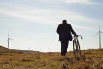 Visão traseira do homem puxando a bicicleta para cima contra o parque eólico — Fotografia de Stock