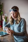 Молода жінка з овочевим соком використовує смартфон на вікні кафе — стокове фото