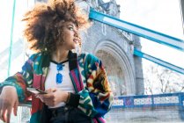 Young girl outdoors holding smartphone when looking away, Tower Bridge in background, London, England, UK — Stock Photo
