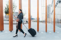 Businesswoman pulling wheeled suitcase and using smartphone outdoors — Stock Photo