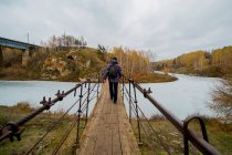 Rear view of young male hiker walking over river footbridge, Kislokan, Evenk, Russia — Stock Photo