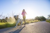 Mujer joven corriendo por carretera rural con perro - foto de stock