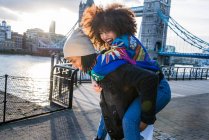Young giving young woman piggyback outdoors, Tower Bridge in background, London, England, UK — Stock Photo