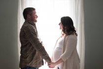 Pregnant woman holding hands with partner, face to face, in front of window — Stock Photo