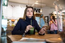 Молода жінка сидить у кафе, тримає смартфон, п'є смузі — стокове фото
