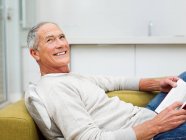 Hombre mayor sentado en un sofá con libro - foto de stock