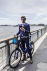 Joven hombre de negocios en bicicleta mirando el teléfono inteligente a lo largo de la orilla del río ciudad - foto de stock