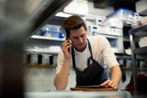 Молодий шеф-кухар читає цифровий планшет і розмовляє на смартфоні на кухні — стокове фото