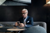 Старший бизнесмен сидит за гостиничным столом, используя сенсорный экран смартфона — стоковое фото