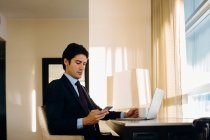 Geschäftsmann schreibt SMS, während er Laptop am Hotelzimmerfenster benutzt — Stockfoto
