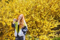 Adolescente debout devant la grande Forsythia jaune, vérifiant son téléphone mobile. — Photo de stock