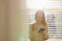 Ragazza adolescente in piedi davanti alla finestra, controllando il suo telefono cellulare. — Foto stock