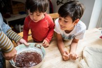 Dois meninos com cabelo preto sentado em uma mesa de cozinha, assando bolo de chocolate. — Fotografia de Stock