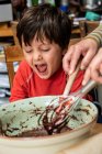 Niño con el pelo negro sentado en una mesa de la cocina, hornear pastel de chocolate. - foto de stock