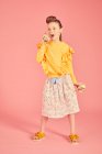 Menina morena usando top amarelo e saia com padrão floral segurando telefone shell mar, no fundo rosa, criança brincalhão — Fotografia de Stock