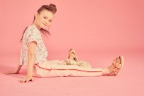 Retrato de chica morena sobre fondo rosa, vistiendo top floral y pantalón rosa pálido sentado en el suelo, mirando a la cámara con sonrisa - foto de stock