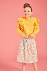 Retrato de chica morena vistiendo top amarillo y falda con patrón floral sobre fondo rosa, mirando a la cámara con feliz dace y sonrisa - foto de stock
