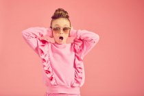 Portrait of brunette girl wearing pink frilly top and sunglasses on pink background, looking at camera with opened mouth, covering ears — Stock Photo