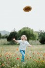 Portrait of young girl with blond hair in a wild flower meadow, throwing straw hat in air. — Stock Photo