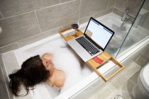 Женщина сидит в ванной, принимает пенную ванну и работает на ноутбуке во время коронавирусного кризиса. — стоковое фото