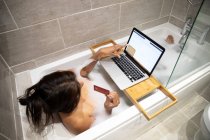 Mujer sentada en la bañera, con baño de espuma y compras en línea en su computadora portátil durante la crisis de Coronavirus. - foto de stock