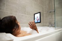 Donna seduta nella vasca da bagno, con bagno di schiuma e utilizzando tablet digitale durante la crisi di Coronavirus. — Foto stock