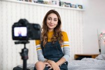 Молода жінка з довгим коричневим волоссям сидить на ліжку, записуючи блог за допомогою цифрової камери . — стокове фото