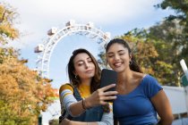 Две молодые женщины с длинными каштановыми волосами стоят в парке возле колеса обозрения, делая селфи с мобильным телефоном. — стоковое фото