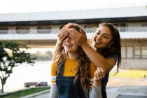 Two young women with long brown hair standing on riverbank, smiling and covering eyes. — Stock Photo